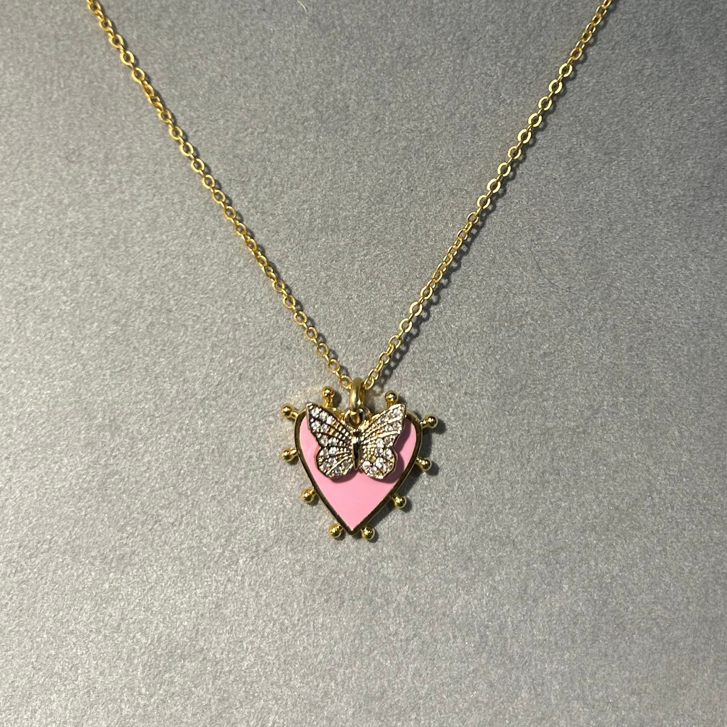 Necklace with a heart-shaped pendant featuring a butterfly design on a gray background, branded 'Pom Di Pom'.