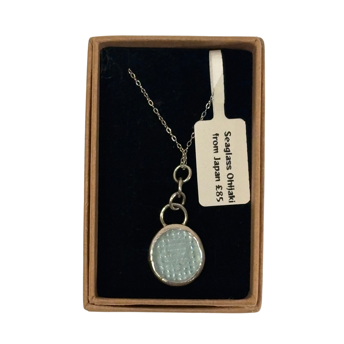 A silver chain necklace with a textured, oval-shaped light blue pendant, presented inside a brown cardboard gift box with a black velvet lining and a paper tag.