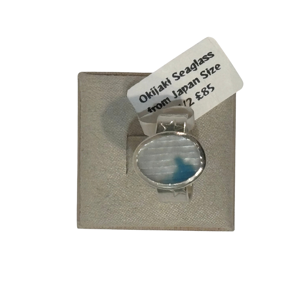 A silver-toned ring with a round bezel containing milky white seaglass with a blue inlay, mounted on a textured beige display card with a white tag reading 'Seaglass from Japan 