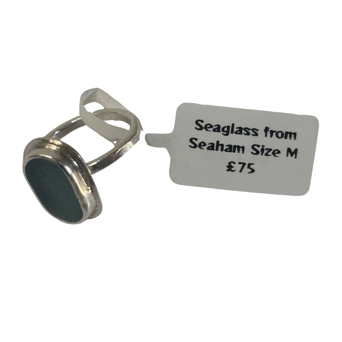 A silver-toned seaglass ring with a dark blue-green oval stone, shown next to a paper tag that reads 'Seaglass from Seaham Size M £75'.