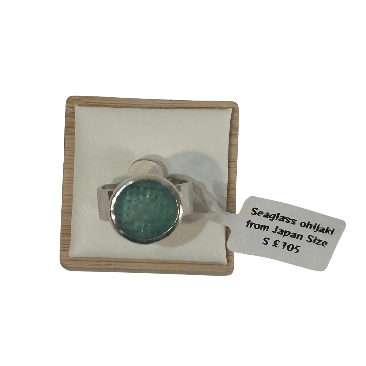 A silver-toned ring with a round textured green seaglass, presented on a cream cushioned wooden display box with a white price tag attached.
