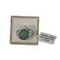 A silver-toned ring with a round textured green seaglass, presented on a cream cushioned wooden display box with a white price tag attached.