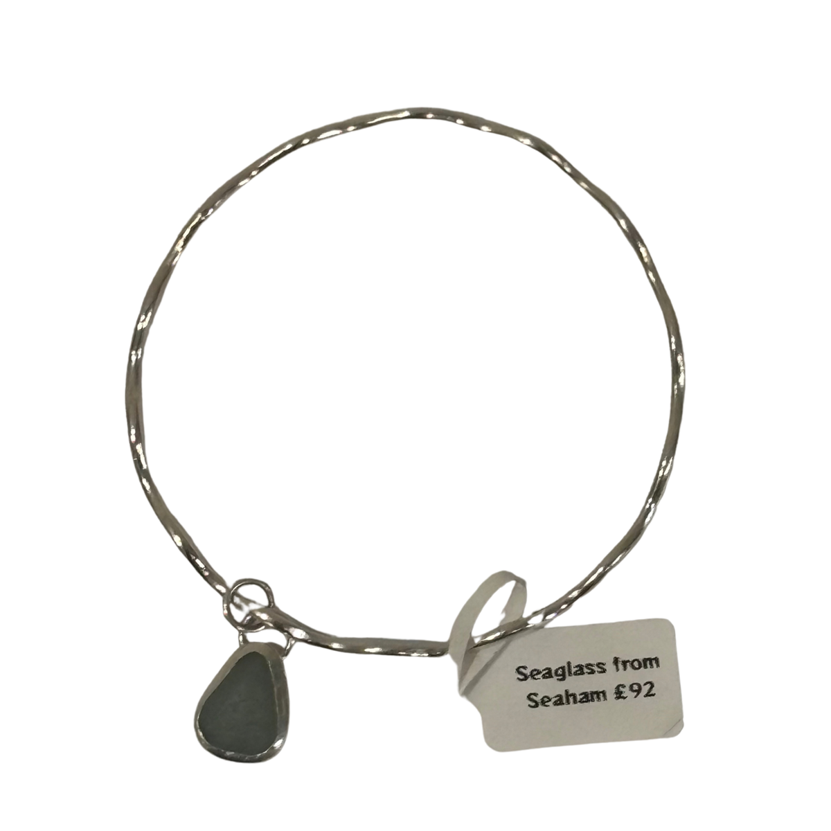 A silver-toned wire bangle bracelet with a small teardrop-shaped seaglass charm and a paper tag that reads 'Seaglass from Seaham £92', shown on a plain white background.