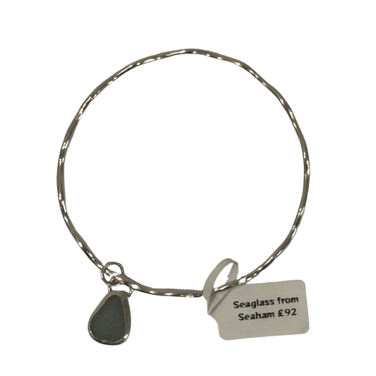 A silver-toned wire bangle bracelet with a small teardrop-shaped seaglass charm and a paper tag that reads 'Seaglass from Seaham £92', shown on a plain white background.