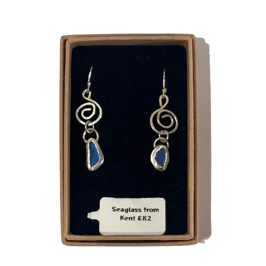 A pair of silver-toned dangle earrings with blue seaglass stones, presented in a brown gift box with a black velvet insert and a price tag reading 'Seaglass from Kent £82'.