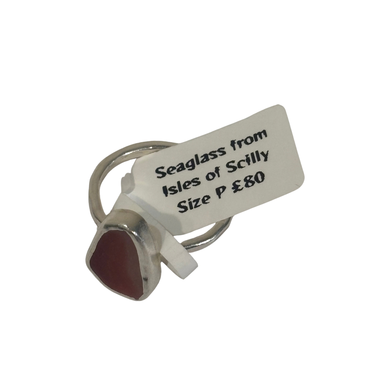 A silver-toned ring with a small, irregularly shaped dark red seaglass inlay, shown with a white paper tag that reads 'Seaglass from Isles of Scilly Size P £80'.