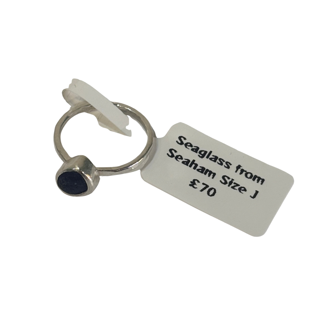 A silver-toned ring with a small oval dark blue stone, attached to a white paper tag that reads 'Seaglass from Seaham Size J £70' on a plain white background.